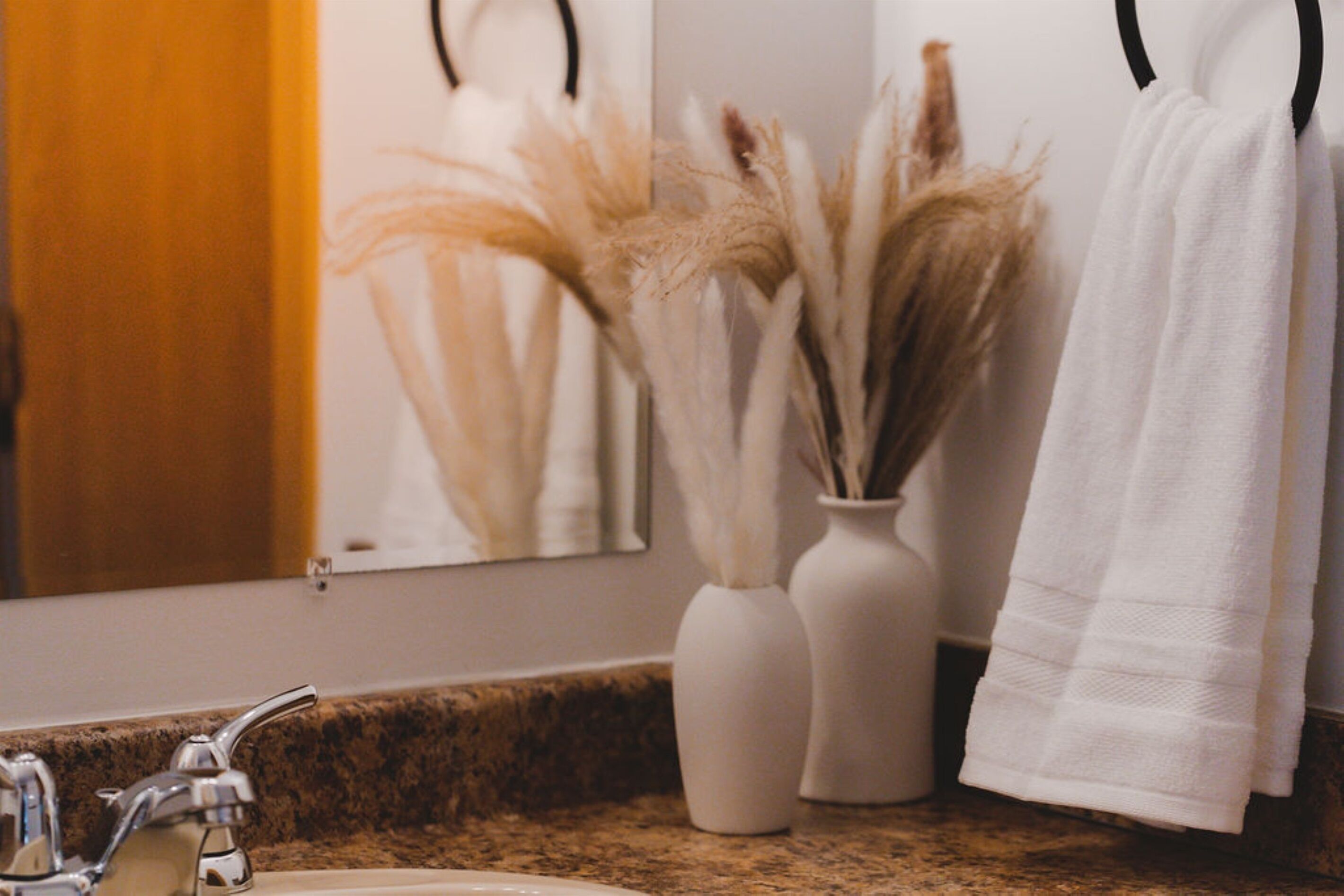 A bathroom with a granite countertop, a white sink, two white vases with dried plants, and a towel hanging on a black towel ring.