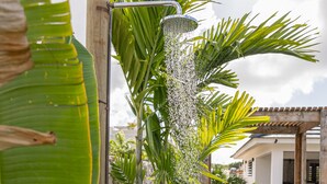 Una ducha exterior moderna con un poste de madera, rodeada de plantas verdes exuberantes y un césped bien cuidado.