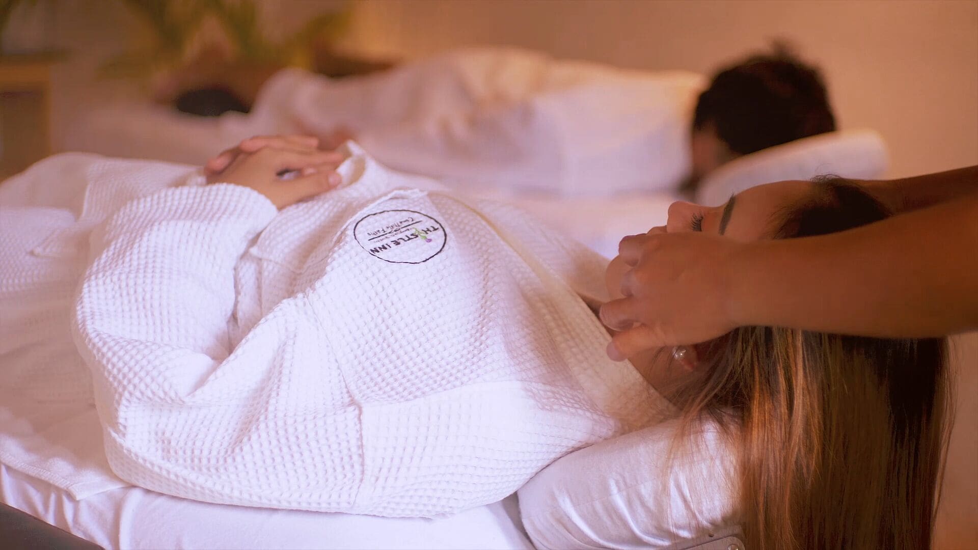 A person lying on a bed in a white robe, with another person attending to them.