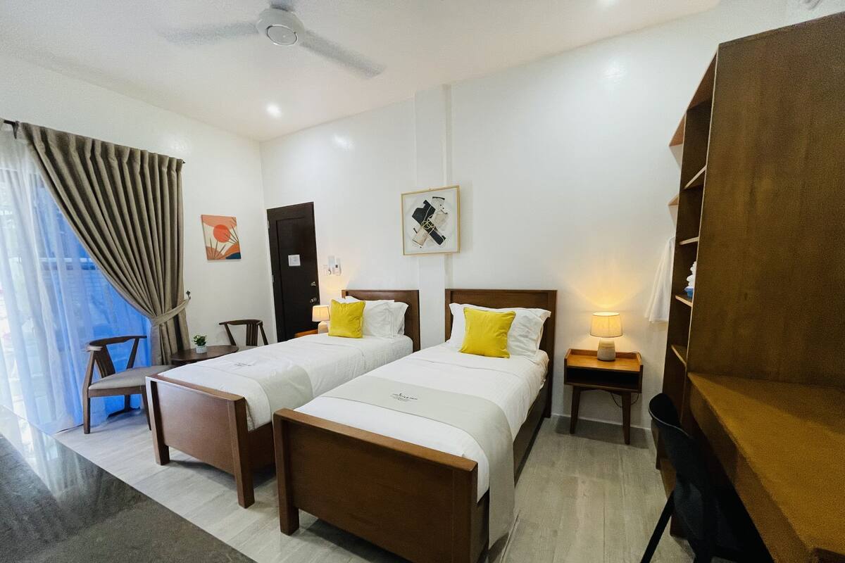 Habitación de hotel con dos camas, un escritorio, una silla, una estantería y un ventilador de techo.