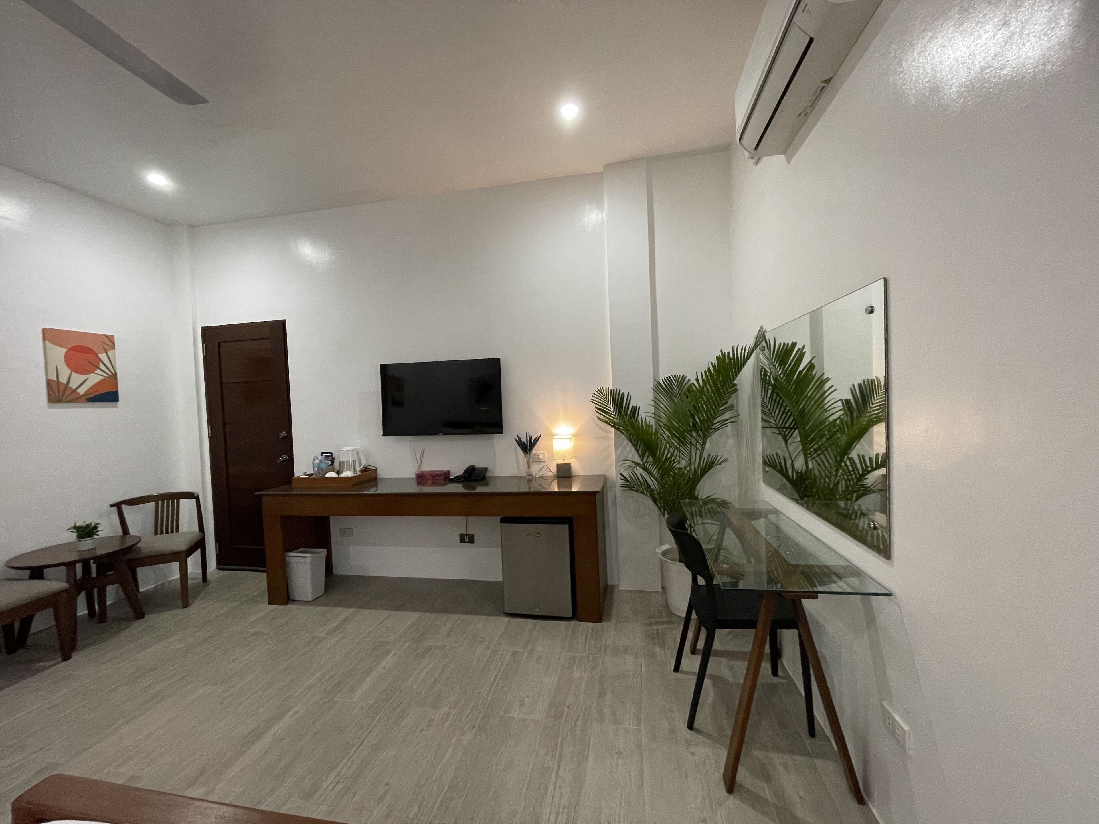 A modern living room with a wooden desk, a flat-screen TV, a small potted plant, and a dining area with wooden chairs and a table.