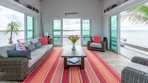 Ein geräumiges Wohnzimmer mit großem Fenster mit Blick auf ein Gewässer, einem Sofa mit gemusterten Kissen, einem Couchtisch und einem Korbstuhl.