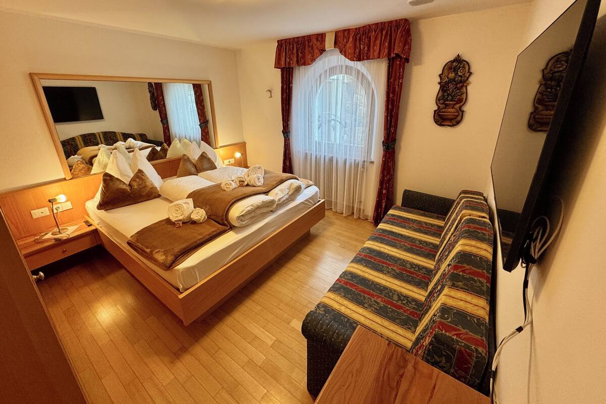 A hotel room with a large bed, a bedside table with a lamp, a sofa, a mirror, and a window with curtains.
