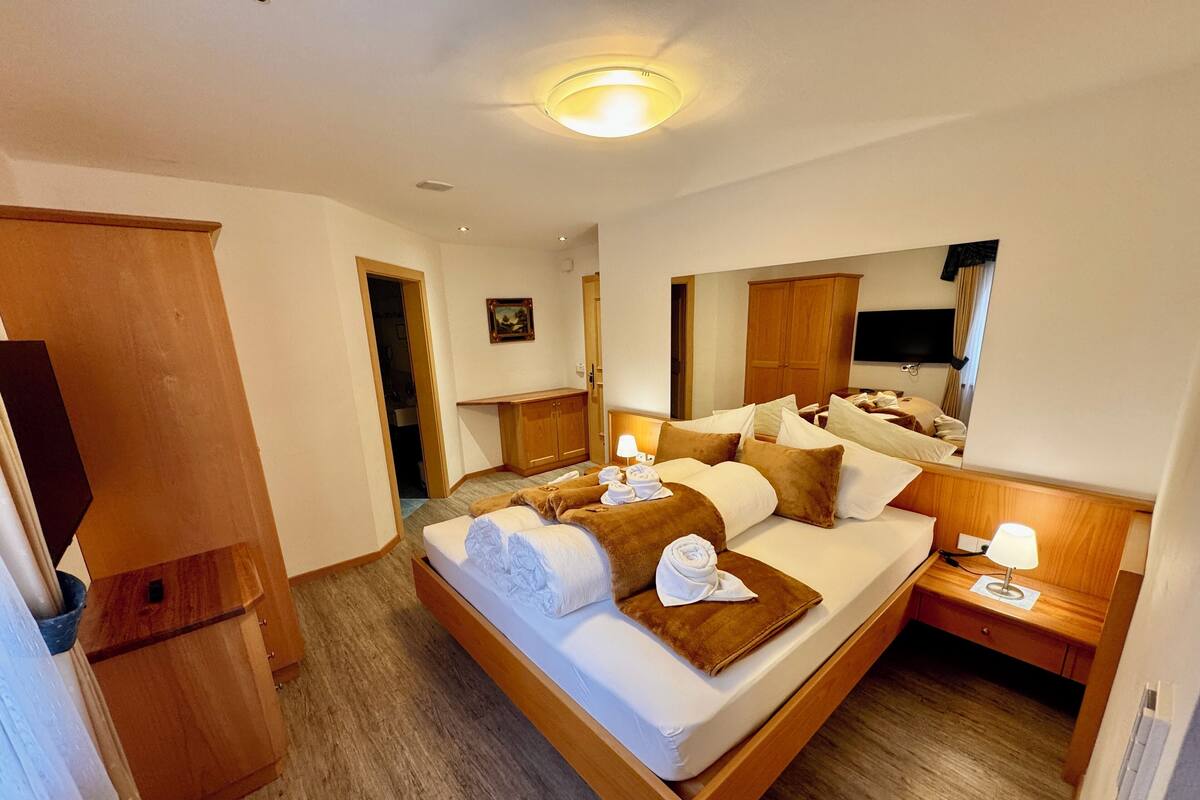 A hotel room with a large bed, wooden headboard, a bedside table with a lamp, a TV mounted on the wall, and a wooden wardrobe.