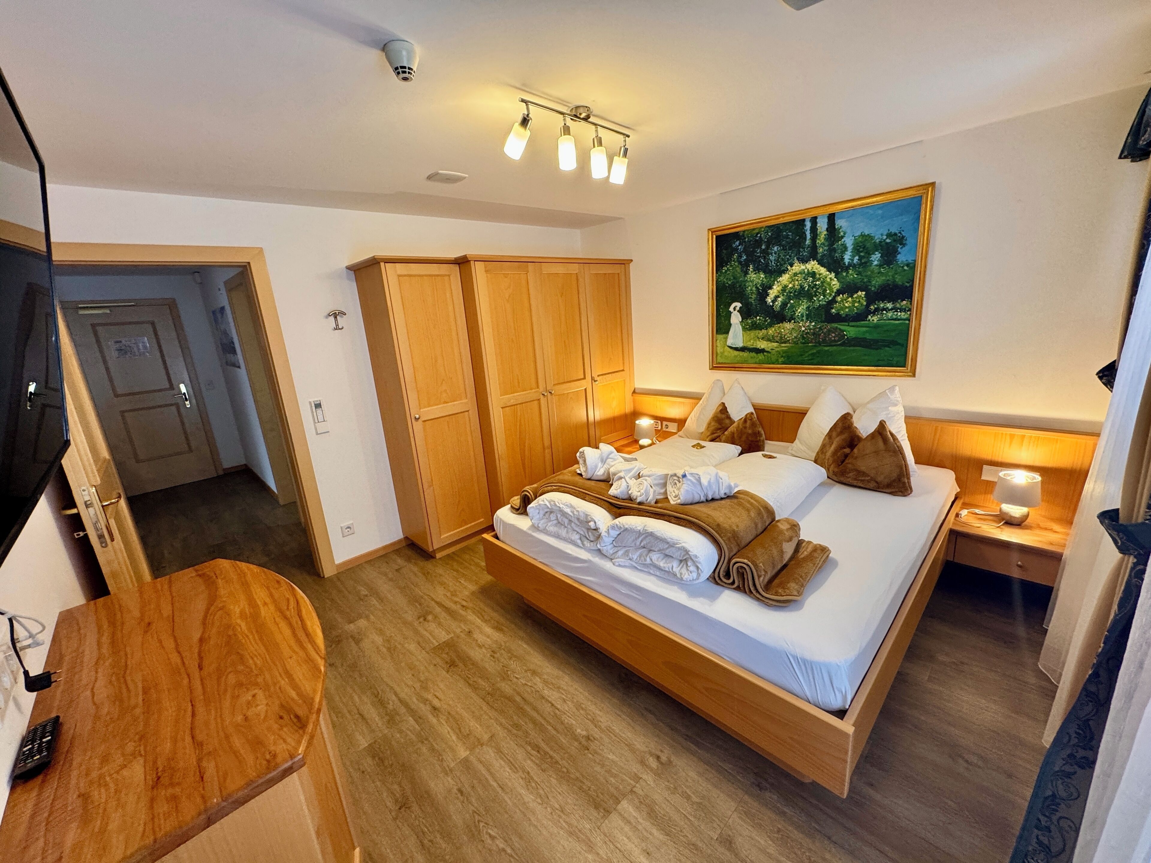 A hotel room with a large bed, wooden wardrobe, and a painting of a garden.