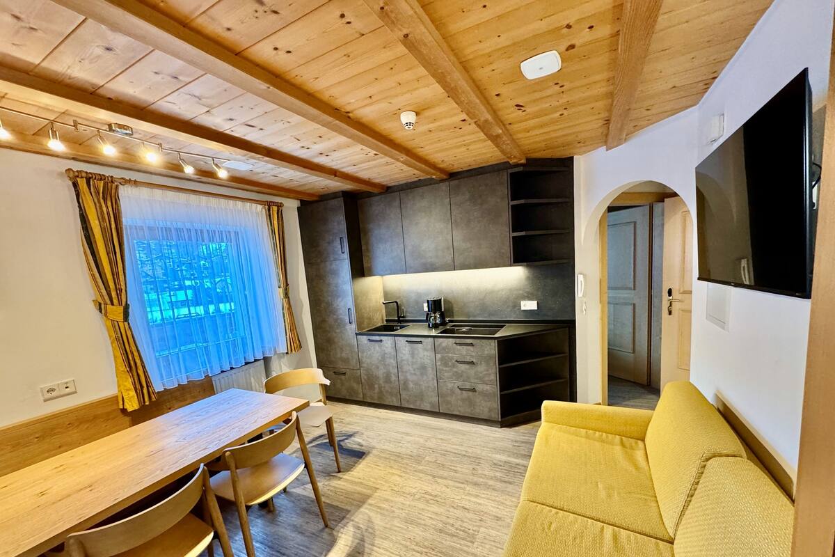 A modern kitchen with dark cabinets and a built-in stove, a dining area with a wooden table and chairs, and a yellow sofa in a cozy living space.