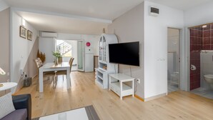 Ein modernes Wohnzimmer mit Flachbildfernseher, einem Essbereich mit Tisch und Stühlen sowie einer Küche mit weißen Schränken und einem Kühlschrank.