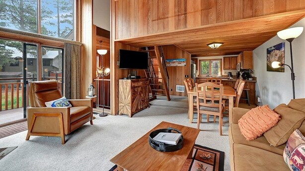 Amplia sala de estar con techo alto, paredes de madera, un gran ventanal, un sofá, una mesa de centro y una zona de cocina con una mesa y sillas para comer.