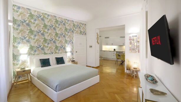 Une chambre avec un lit, une table de chevet, une commode, un téléviseur fixé au mur et, au fond, un coin cuisine.