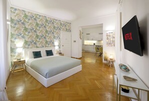 Een slaapkamer met een bed, nachtkastje, wandtelevisie en een keuken op de achtergrond.