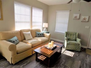 A living room with a beige sofa, a patterned armchair, a coffee table with candles, and a lamp on a side table. - The Aviator's Landing (Pensacola)