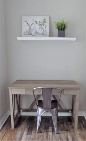 A desk with a chair, a shelf with a painting and a plant, and a wooden floor. - The Aviator's Landing (Pensacola)