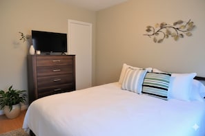 A bedroom with a bed, a dresser with a flat-screen TV, a potted plant, and a wall art piece.