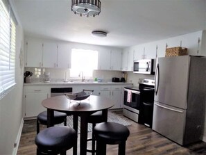A modern kitchen with white cabinets, a stainless steel refrigerator, a microwave, and a dining area with a round table and four stools. - The Aviator's Landing (Pensacola)