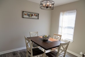 A dining area with a wooden table, white chairs, a chandelier, and a window with blinds.