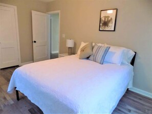 A bedroom with a bed, a door, a nightstand, and a wall-mounted lamp. - The Aviator's Landing (Pensacola)