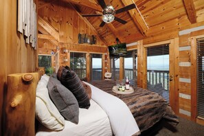 A wooden cabin bedroom with a bed, pillows, a ceiling fan, and a mounted television.