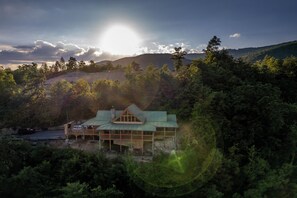 A wooden cabin with a green roof surrounded by dense forest and hills.