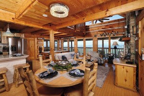 A wooden cabin interior with a dining area, kitchen, and seating by a large window.