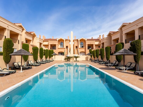 A pool area with lounge chairs and umbrellas, surrounded by buildings with a symmetrical architectural design.