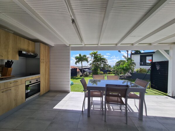 A modern kitchen with wooden cabinets and a built-in oven, leading to an outdoor dining area with a glass table and chairs.