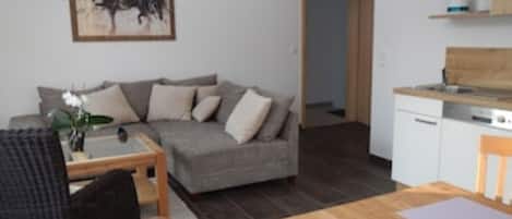 Una sala de estar con un sofá gris, una mesa de centro de madera y una mesa de comedor con individuales. Se observa una zona de cocina con lavavajillas y encimera de madera. En la pared, hay un cuadro enmarcado.