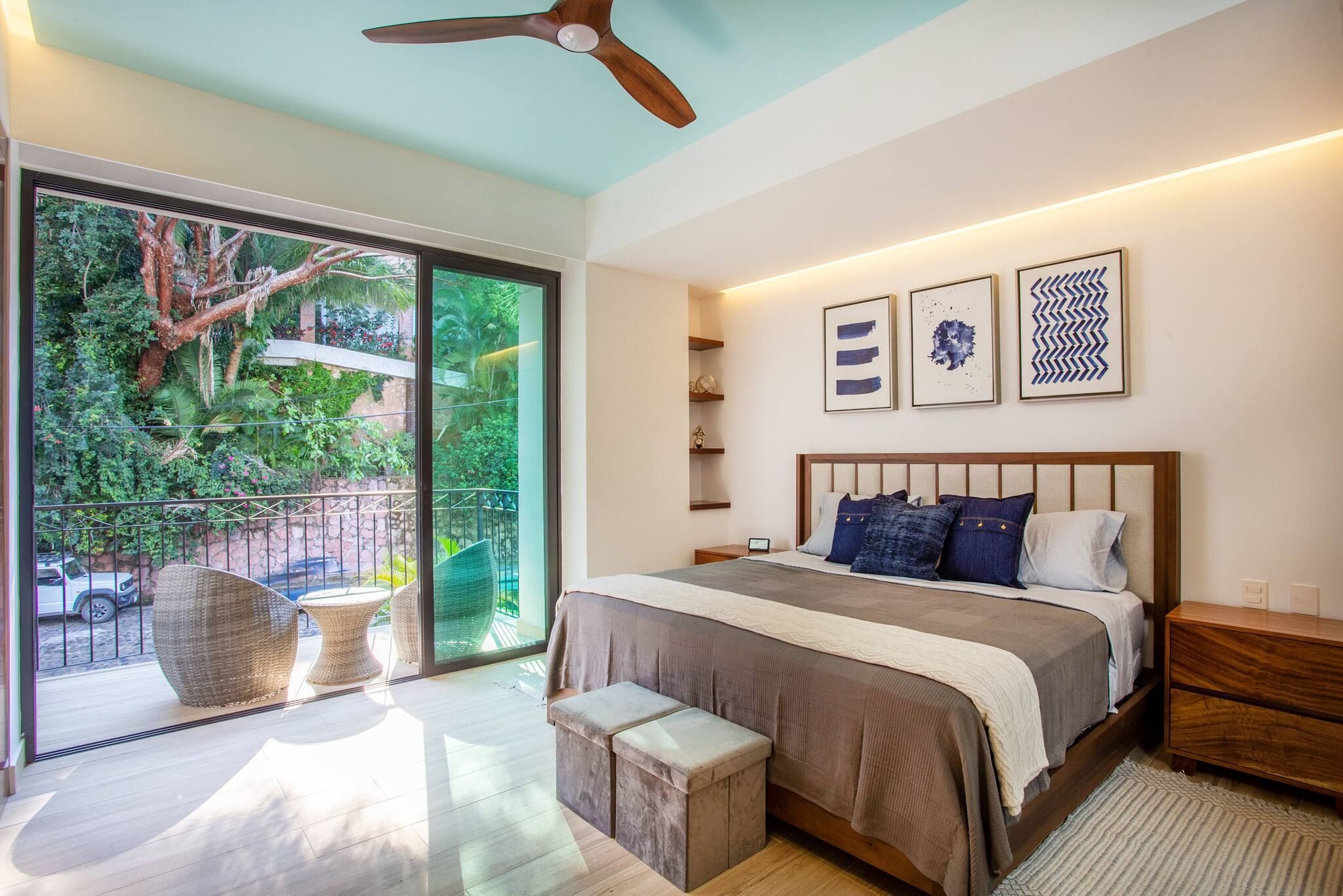 A bedroom with a large bed, a ceiling fan, a sliding glass door leading to a balcony, and artwork on the wall.