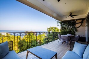 Ein Balkon mit Tisch und Stühlen, einem Deckenventilator und Blick auf das Meer und Grünflächen.