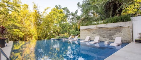 An infinity pool with lounge chairs, surrounded by lush greenery and a stone wall.