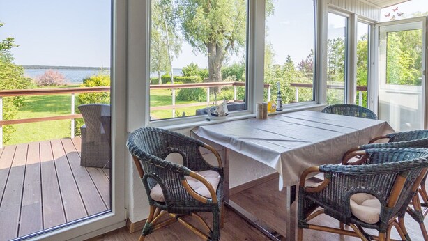 Une terrasse couverte, dotée d’une table et de chaises en osier, qui offre une vue sur une zone herbeuse et un plan d’eau.