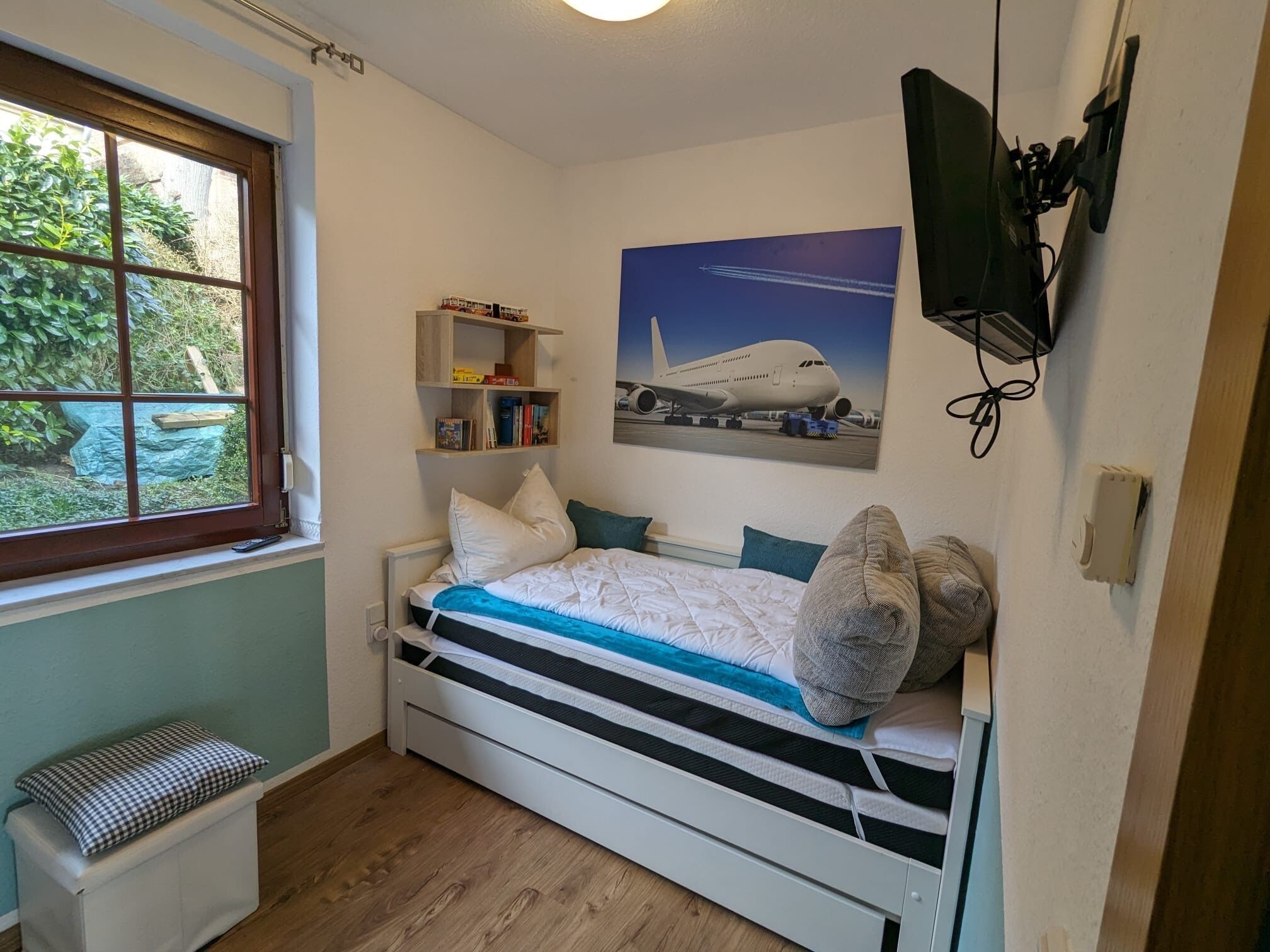 Ein Schlafzimmer mit Bett, Fenster, Bücherregal und Wandfernseher.