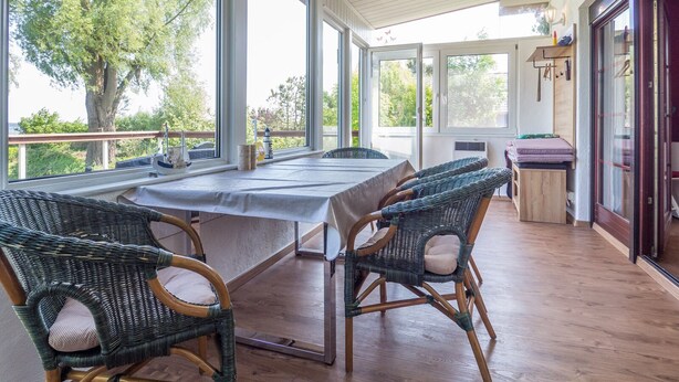 A covered patio with a dining table and wicker chairs, large windows, and a view of trees. - Vacation home Vesteralen in Breege on the Bodden (Breege)