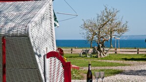 Ein Korbstuhl mit rotem Träger, eine Weinflasche und zwei Gläser auf einem Tisch, mit Blick auf das Meer und einem Baum im Hintergrund.