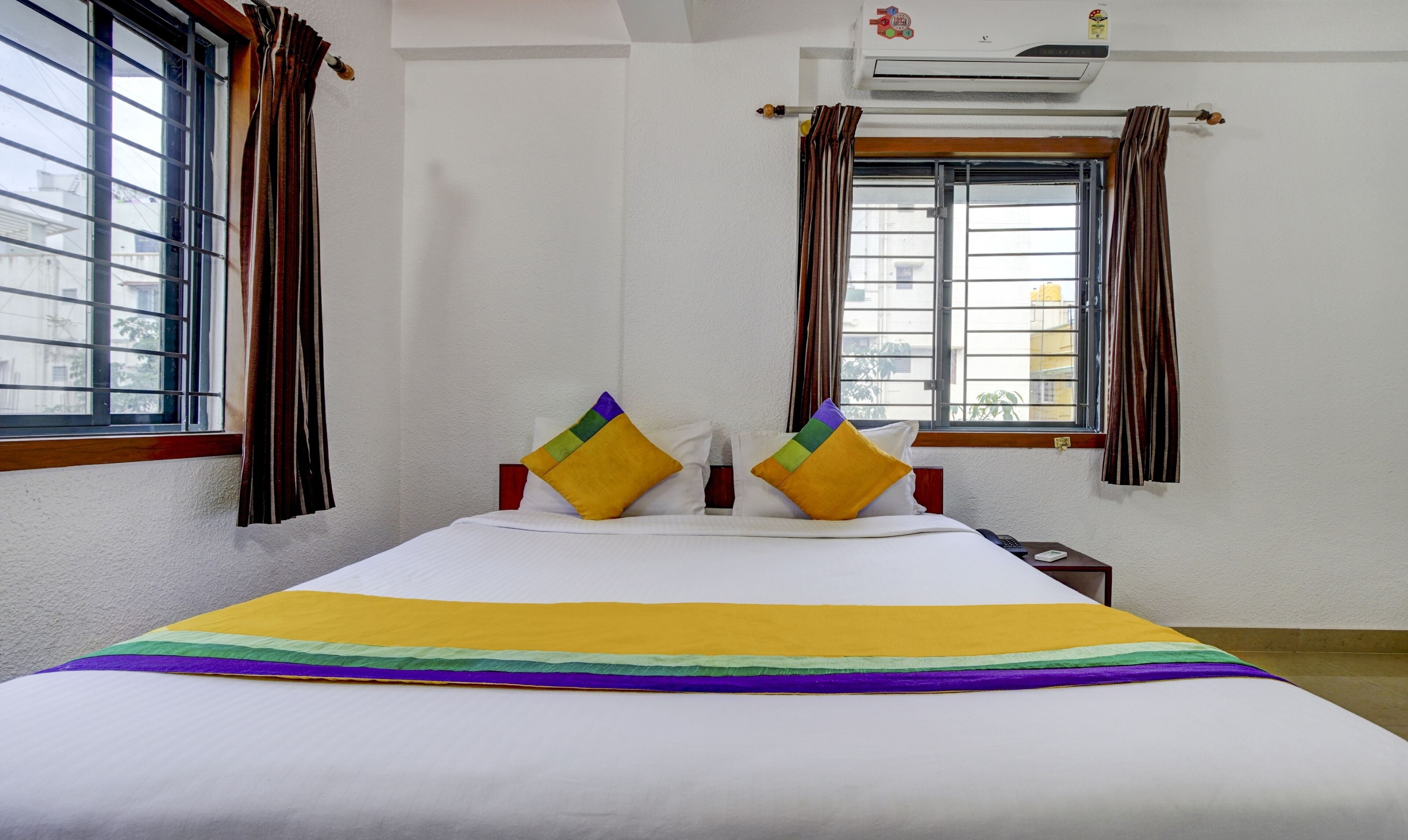 Habitación de hotel con cama, dos ventanas con cortinas, un aire acondicionado y vistas a edificios.