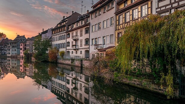 Een rij traditionele Europese gebouwen langs een rustige rivier bij zonsondergang.