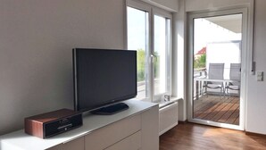 Une chambre moderne avec un téléviseur à écran plat posé sur un meuble blanc, de grandes fenêtres et une vue sur l’extérieur.