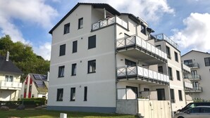 Un immeuble résidentiel à plusieurs étages, doté de balcons, d’une allée pavée et d’une voiture garée devant.
