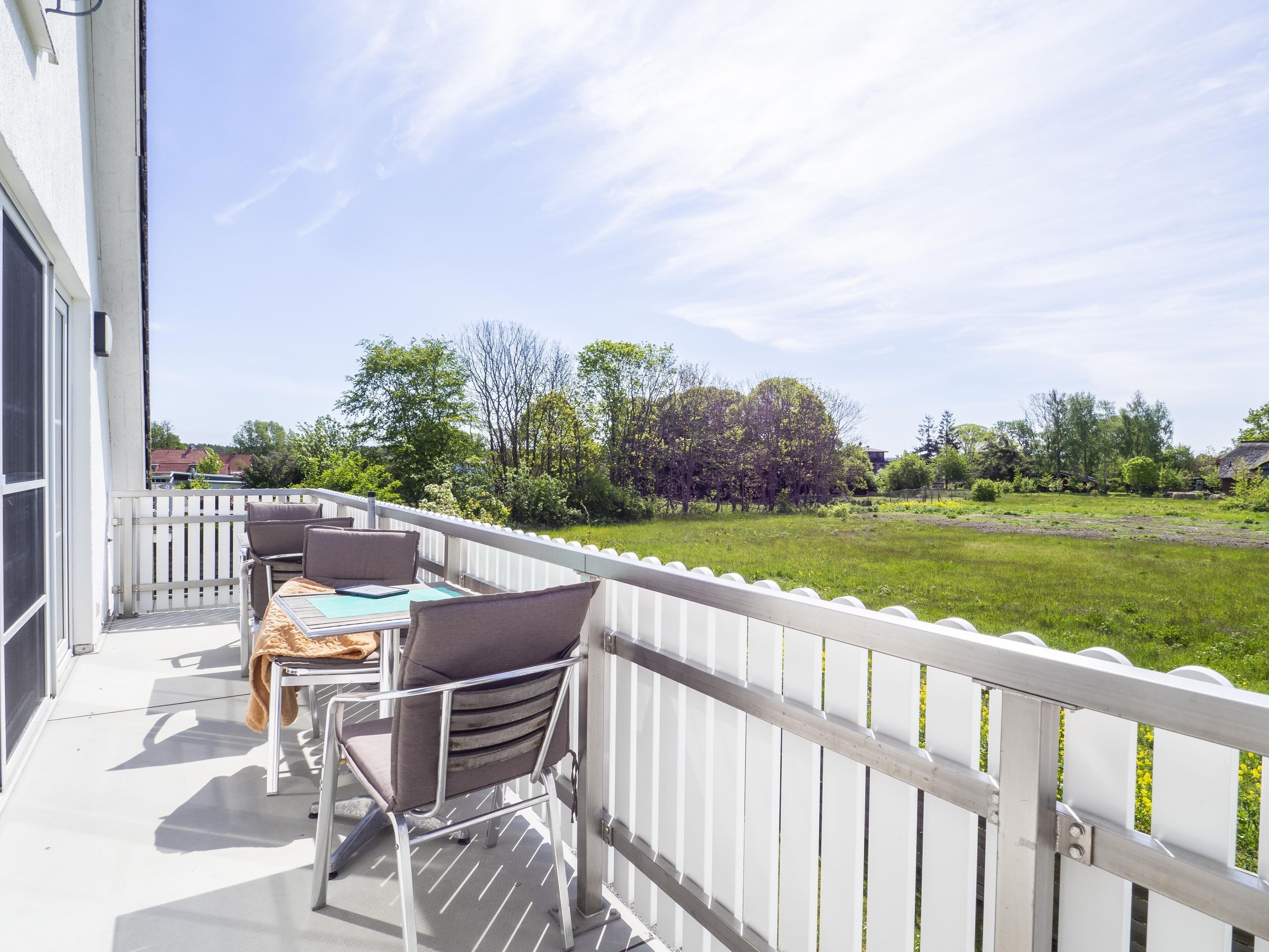 Ein Balkon mit Tisch und Stühlen, der auf eine Wiese mit Bäumen und ein Gebäude im Hintergrund blickt.