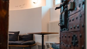 A room with a wooden bench, a small table, and a radiator. There is a sign on the wall that says "THIS WAY OUT!" and a large, ornate door. - Bogentrakt - Hostel (Chur)