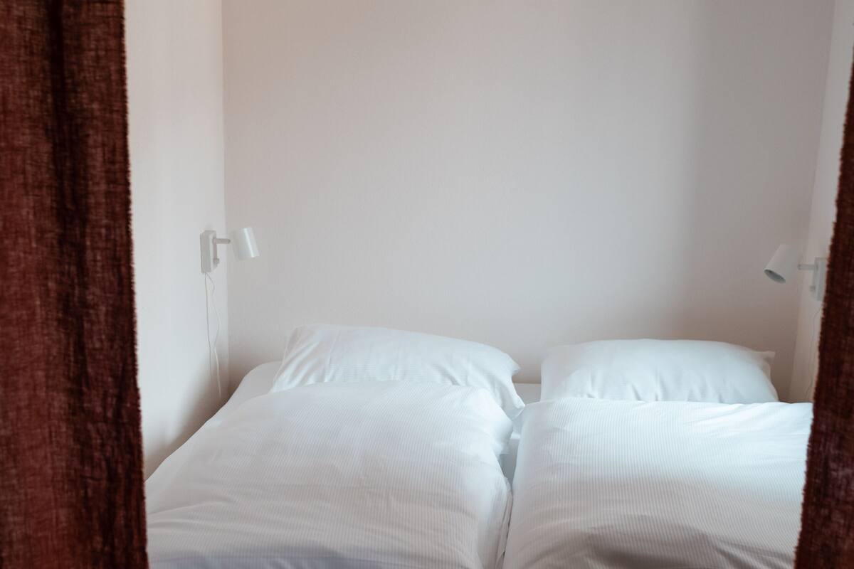 A single bed with white linens in a room with white walls and a brown curtain.