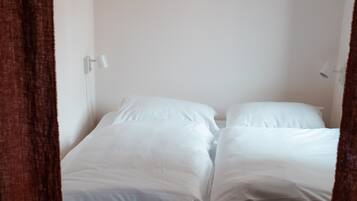 A single bed with white linens in a room with white walls and a brown curtain.