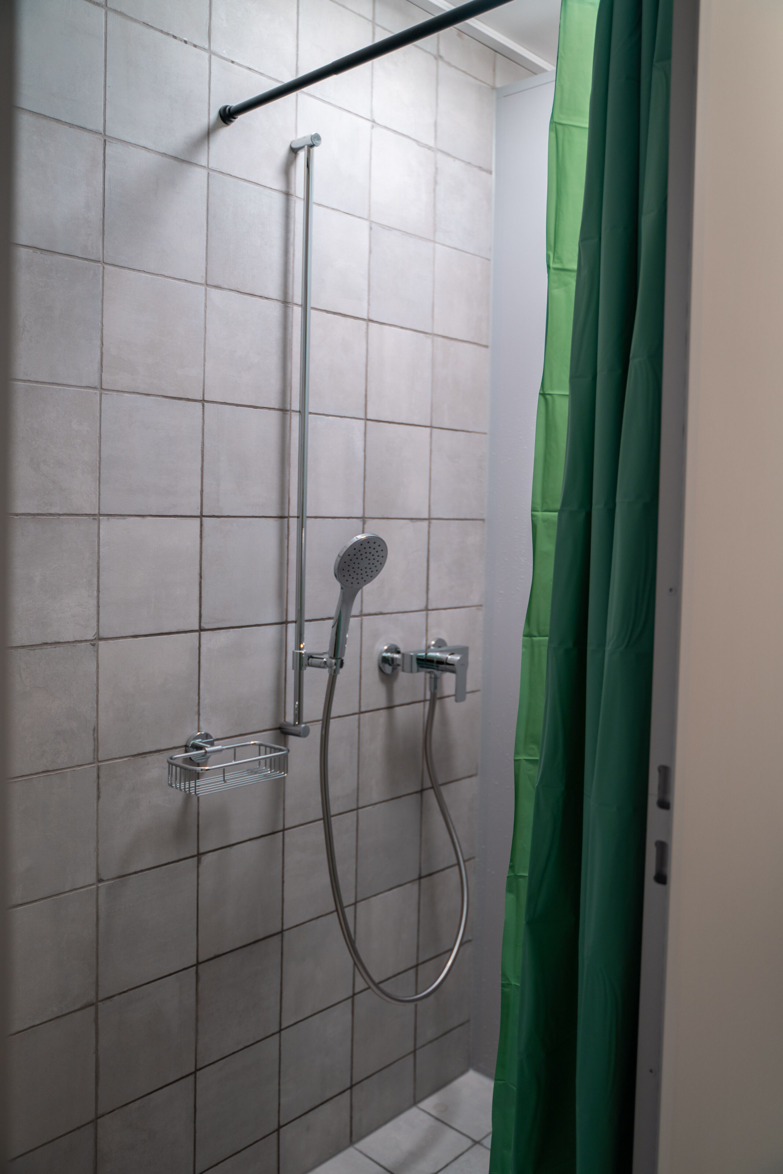 Un área de ducha con cortina verde, cabezal de ducha de mano y grifo instalado en la pared.