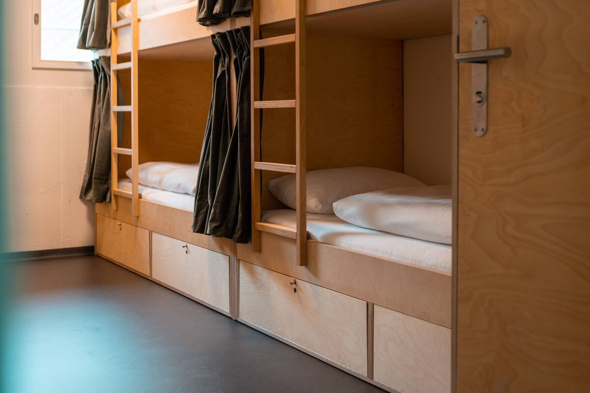 A bunk bed room with a wooden bunk bed, a small shelf, and a door with a number plate.