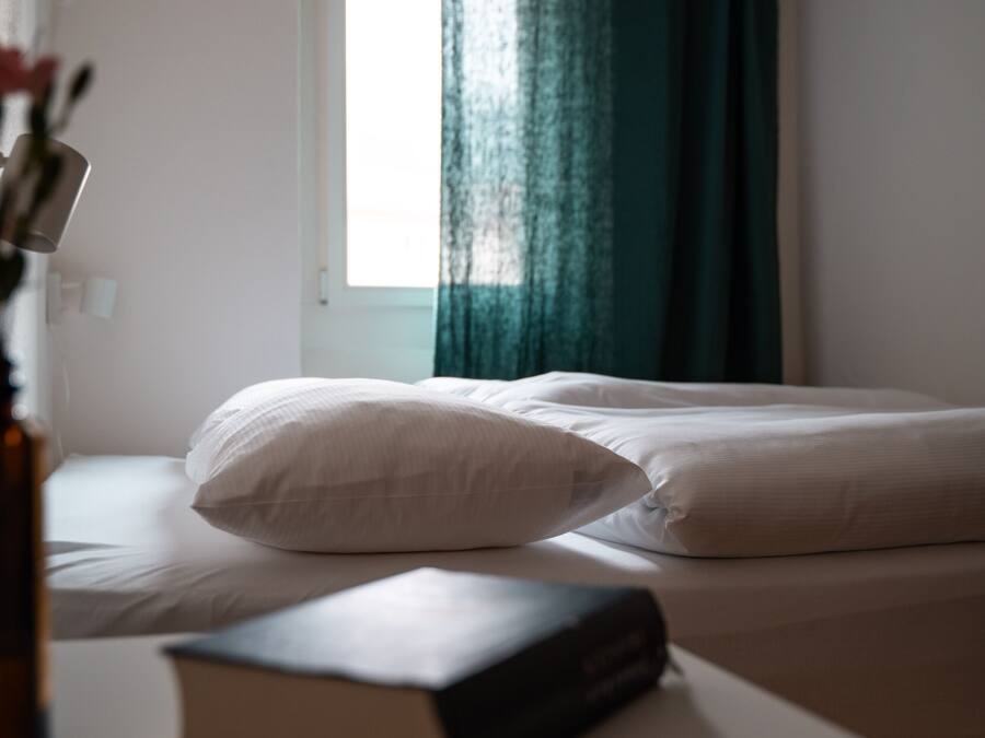 Un dormitorio con cama, una ventana con cortinas y un libro sobre la mesita de noche.