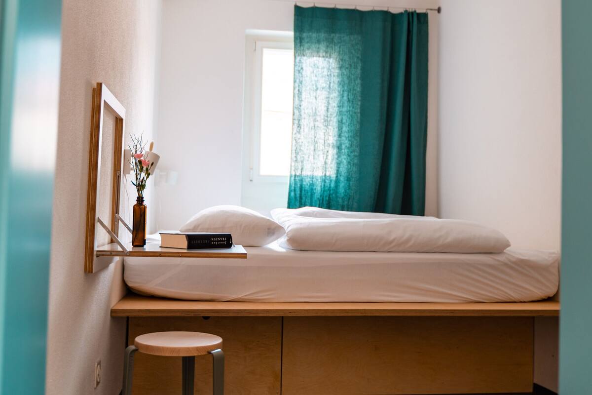 A bedroom with a bed, a small stool, a window with curtains, and a hanging light fixture.