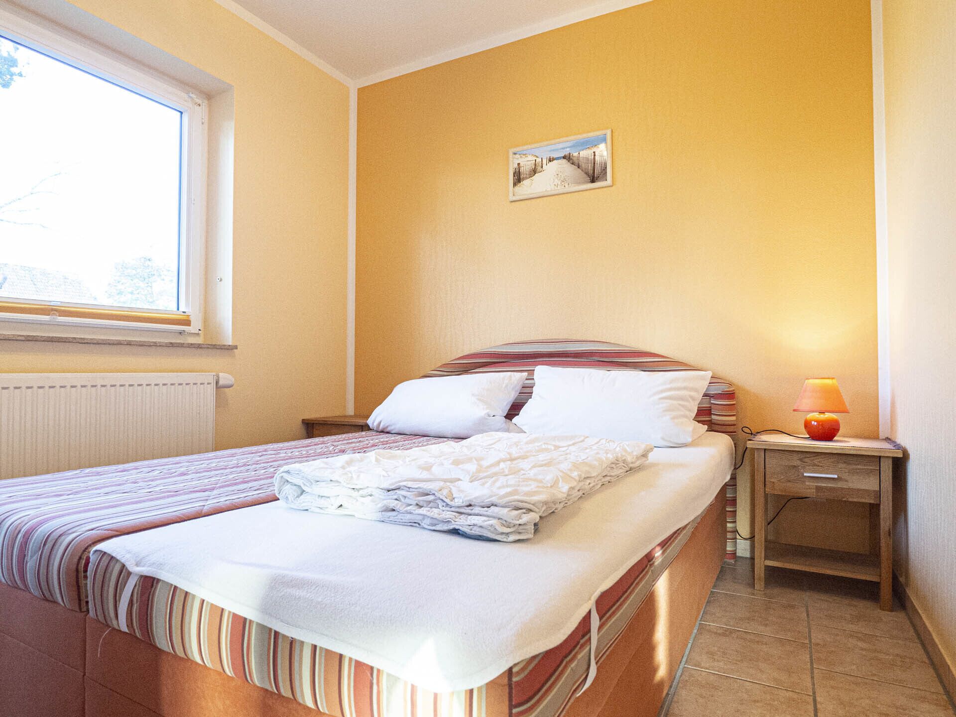 Una cama bien hecha con ropa de cama a rayas, una lámpara de noche y un cuadro en la pared.