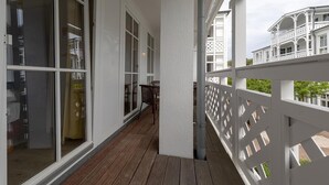 Ein überdachter Balkon mit Holzboden, weißen Geländern und Blick auf ein weißes Gebäude mit zahlreichen Fenstern und Balkonen.
