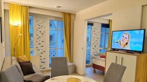 Ein Wohnzimmer mit einer Couch, Sesseln, einem Couchtisch, einem Fernseher und einem Balkon mit gelben Vorhängen.