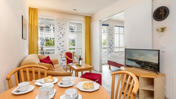 Ein gemütliches Wohnzimmer mit Esstisch, gedeckt für ein Frühstück, einem Fernseher und Blick auf den Balkon.