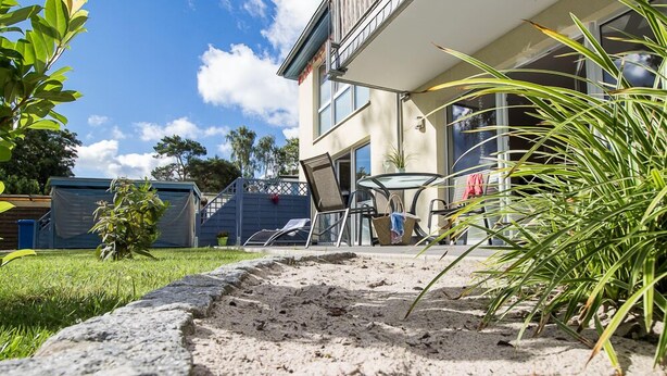 Pihalla on terassialue, jolta kivipolku johtaa lasipöydän ja -tuolien luo. Alueen ympärillä on vehreää kasvillisuutta ja suuret ikkunat omaava rakennus.